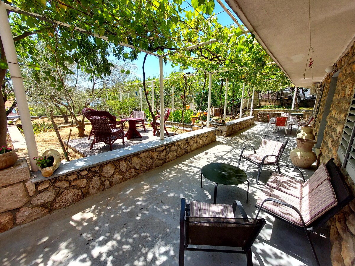 A spacious outdoor area presents a stone terrace shaded by greenery. Several seating options include woven chairs and a table, providing a relaxing space. The garden features various plants and furniture, creating an inviting atmosphere for outdoor gatherings.