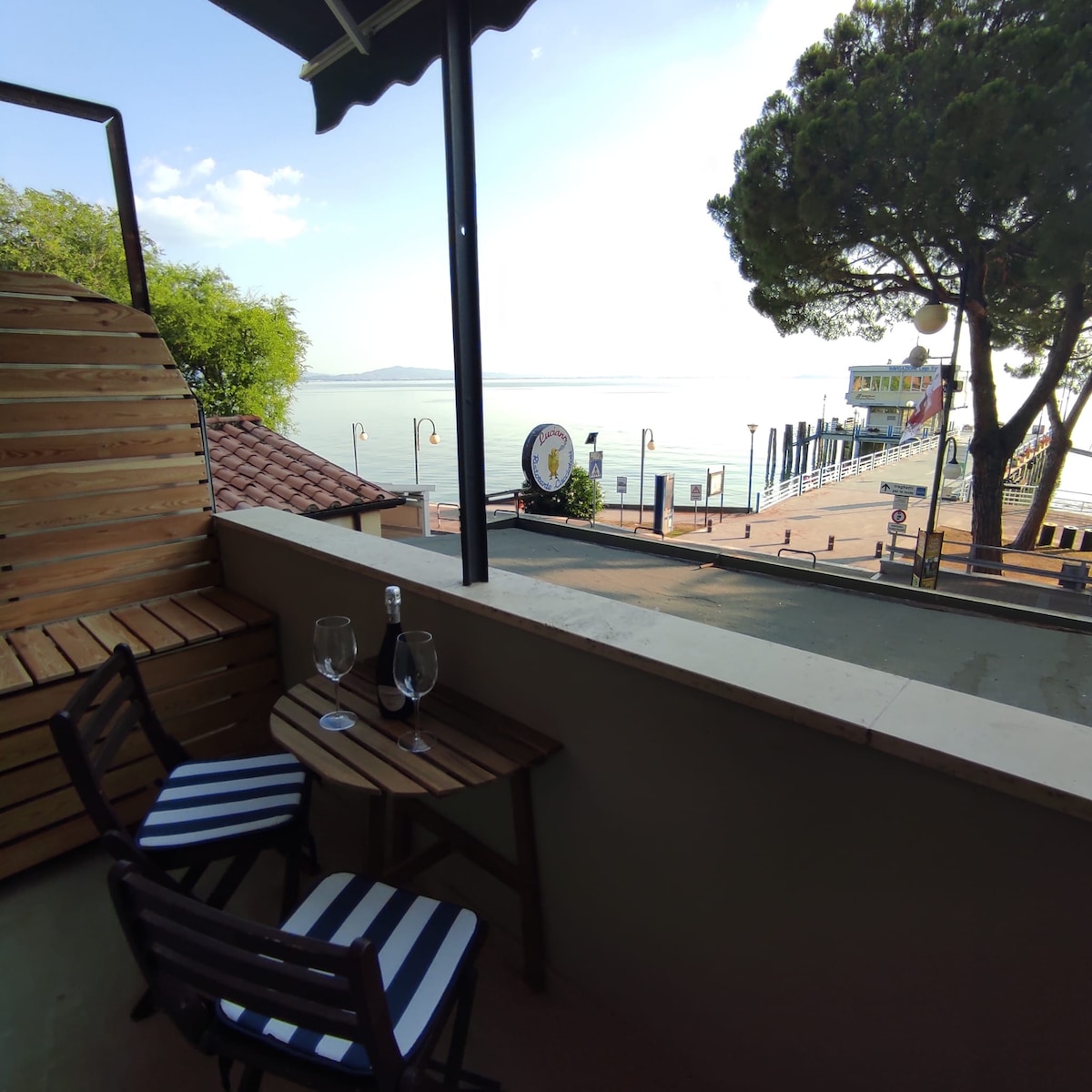 A balcony space overlooks the lake, featuring a small table with two striped chairs. A bottle and glasses are set on the table, while the scenic view includes a nearby ferry dock and a pathway along the water's edge.