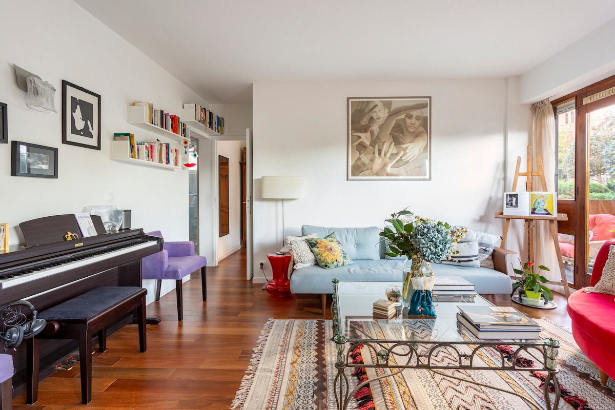 A stylish living area features a mix of modern and classic furnishings. A grand piano is positioned beside a vibrant blue sofa, complemented by decorative pillows. A glass coffee table sits atop a patterned rug, while large windows provide natural light and views of the outdoor space.