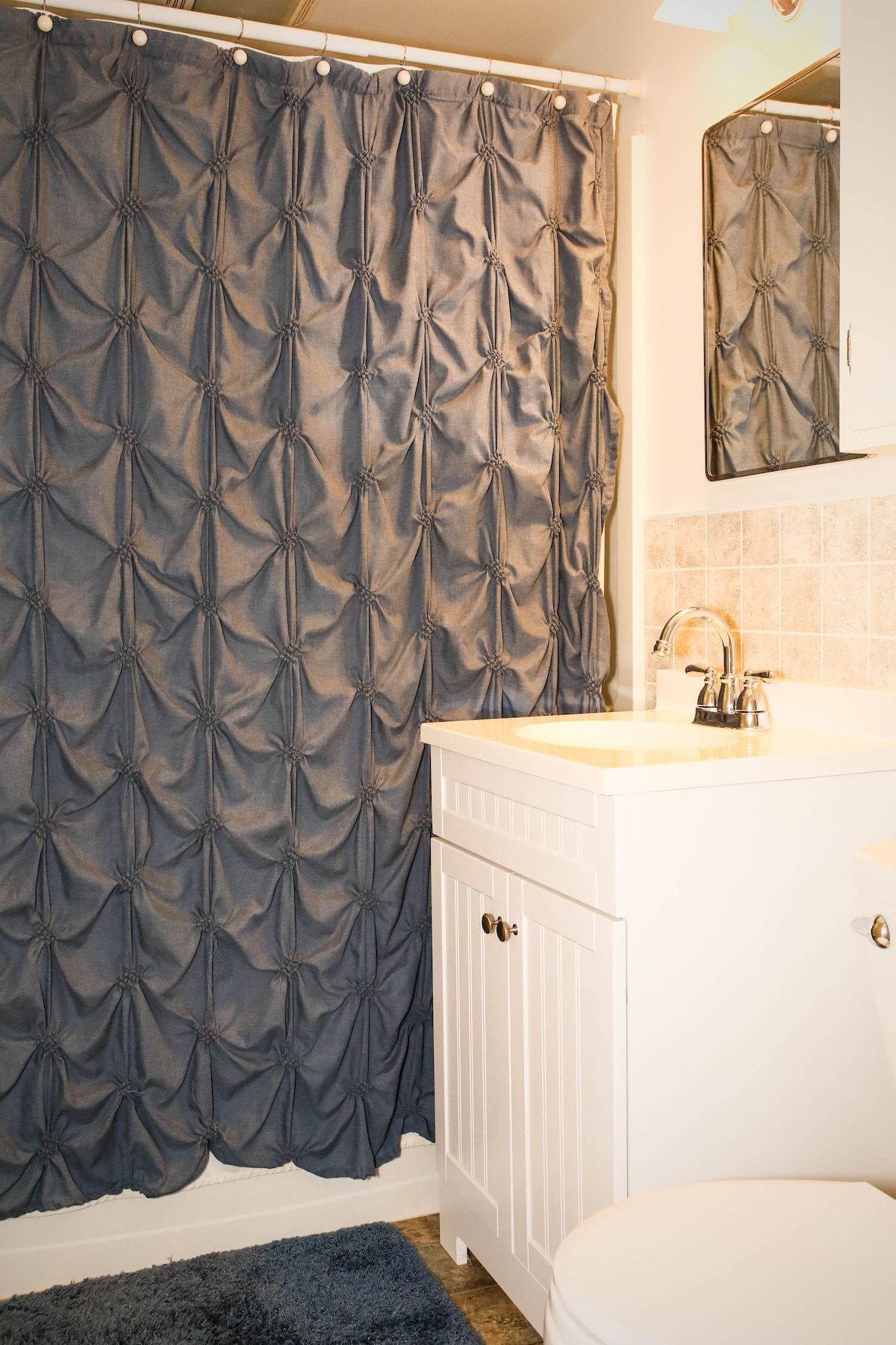 A neatly arranged bathroom features a white vanity with a chrome faucet and a wall-mounted mirror above. A textured navy shower curtain, styled with tufted designs, hangs in front of the bathtub. Light-colored tiles line the walls, enhancing the bright atmosphere.