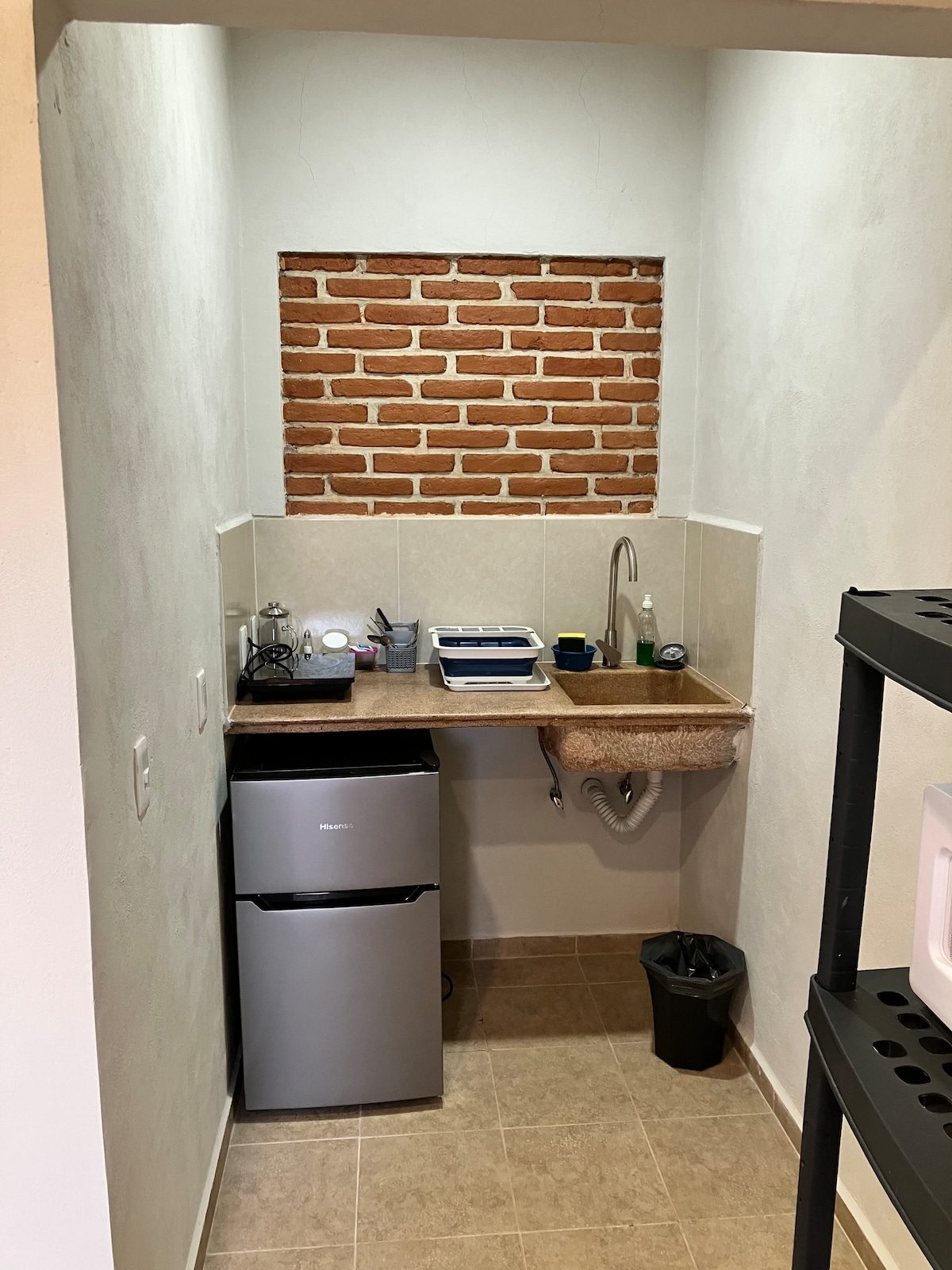 A compact kitchenette is featured, showcasing a small refrigerator, a sink, and a countertop with essential kitchenware. The wall displays a section of exposed brick, adding texture to the space. The flooring is tiled in neutral tones.