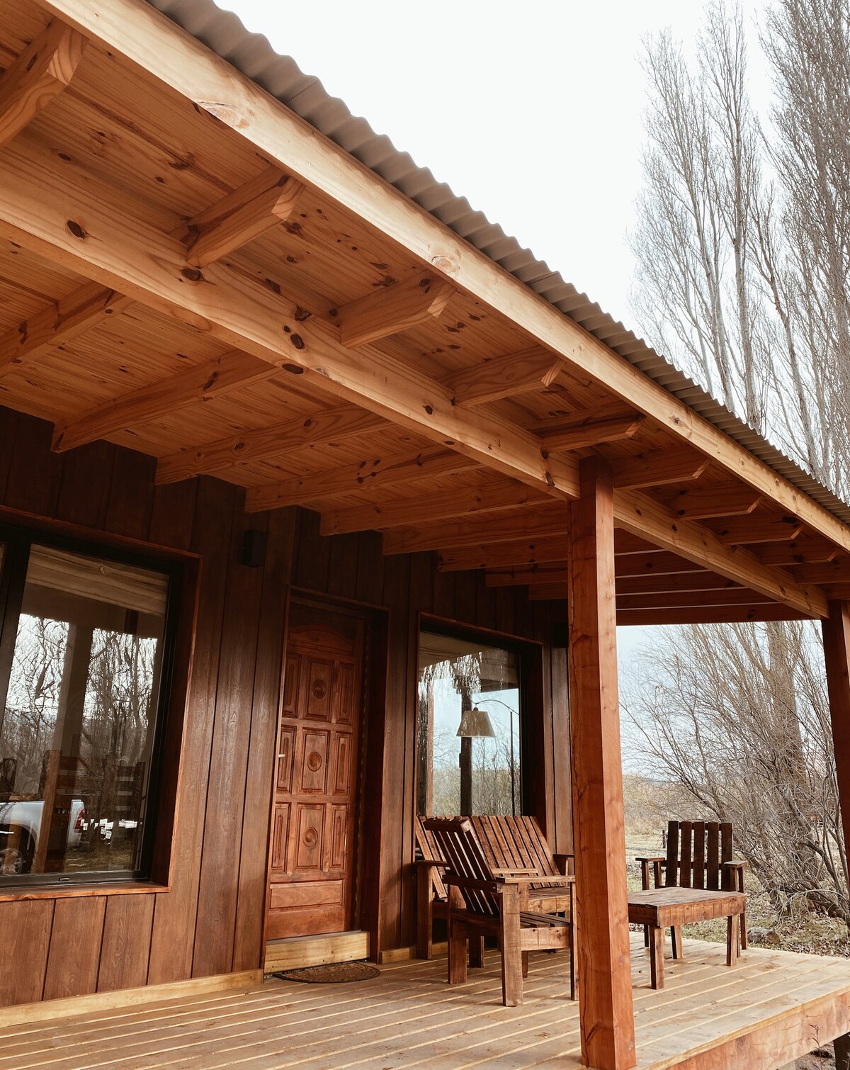 A rustic wooden porch features a solid door, with large windows on either side. Two chairs made of slatted wood are positioned on the deck, inviting relaxation. Tall, slender trees are visible in the background, enhancing the natural setting.