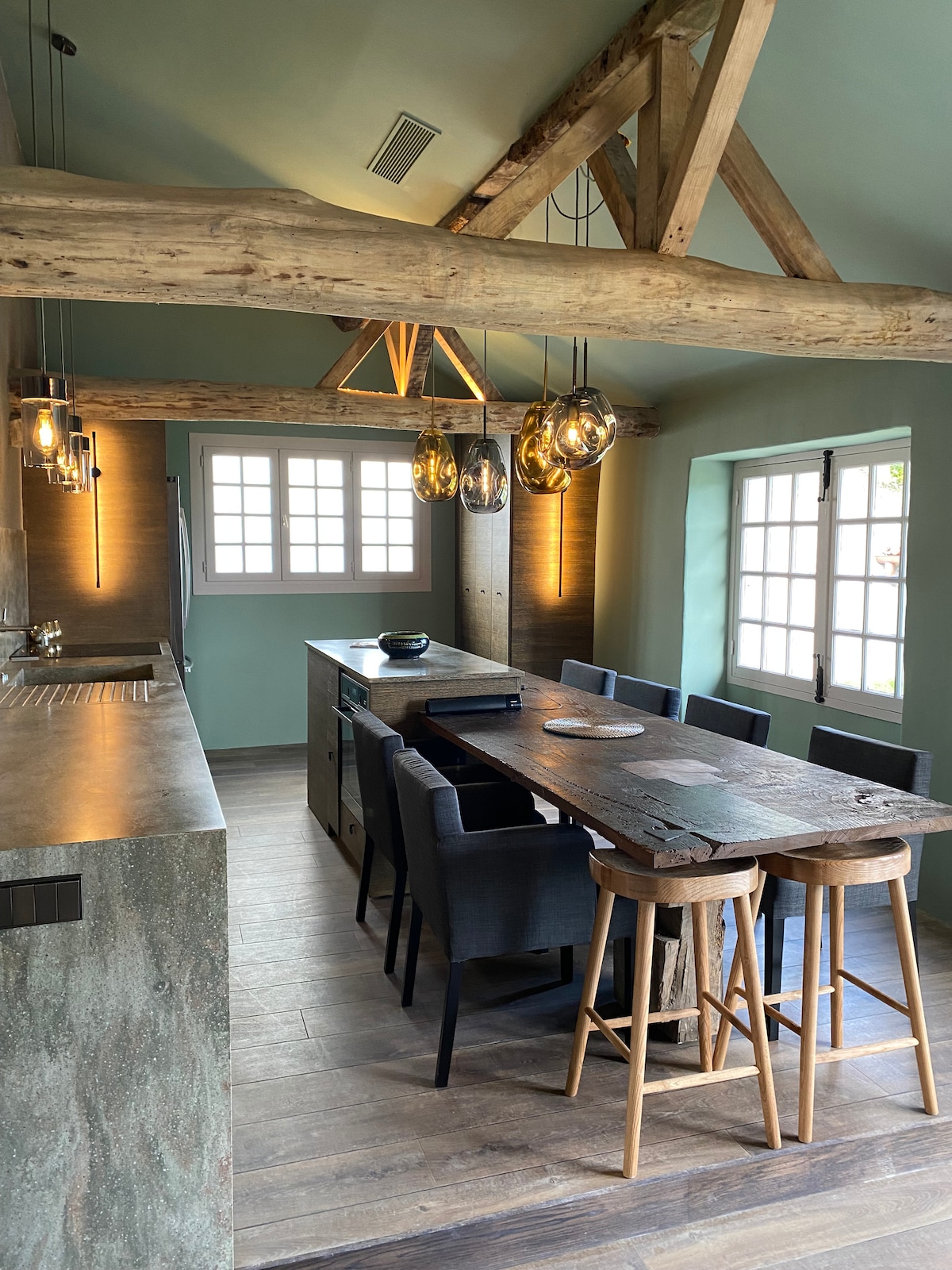 An open-plan kitchen and dining area features a large wooden table surrounded by dark chairs. Pendant lights hang from the ceiling, illuminating the space. Natural materials like exposed wooden beams and stone countertops are prominent. Large windows provide an abundance of natural light.
