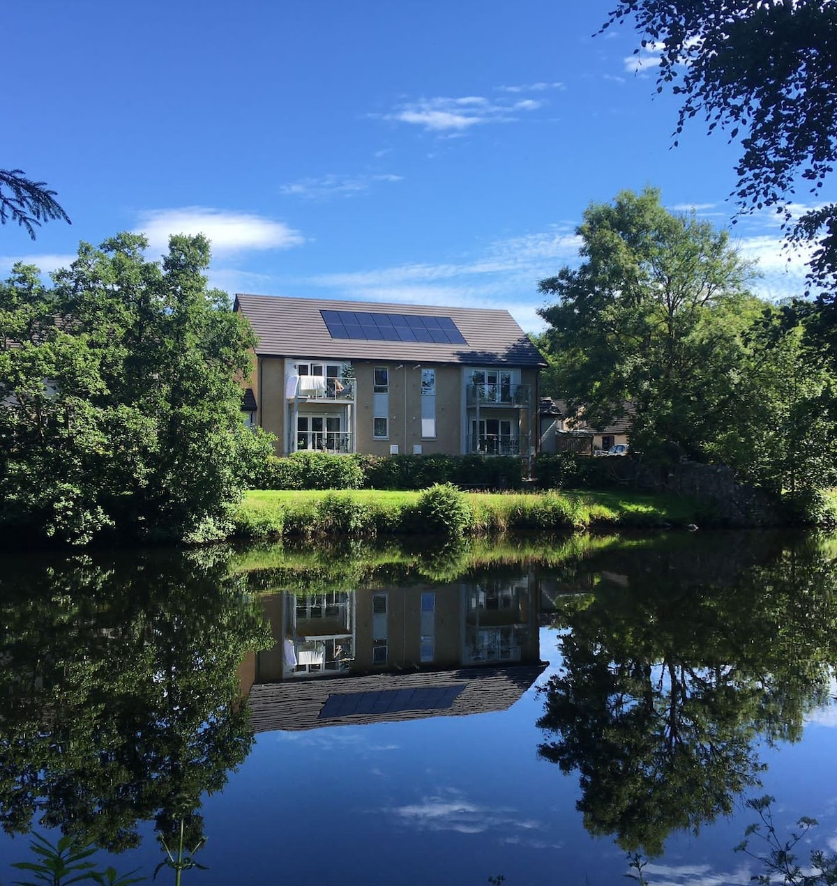 A modern holiday home is depicted, situated by the River Teith. The building features large windows and solar panels on the roof. Lush greenery surrounds the property, creating a serene atmosphere. The reflection of the home can be seen on the calm water, enhancing the tranquil setting.