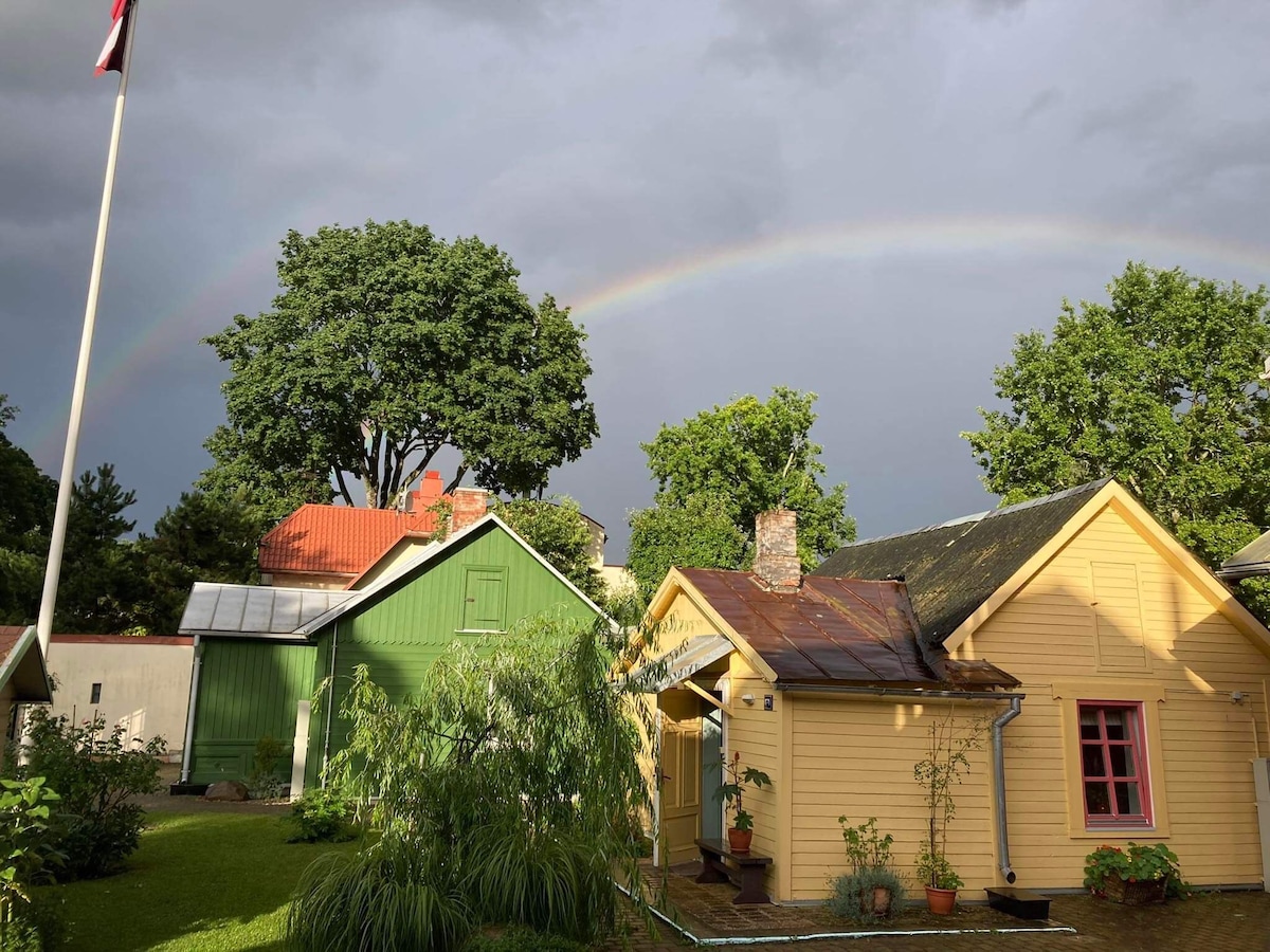A charming yellow house with a wooden exterior is framed by lush greenery and tall trees. A green house and a red-roofed building are visible in the background, with a rainbow arching across the cloudy sky, adding color to the serene setting.