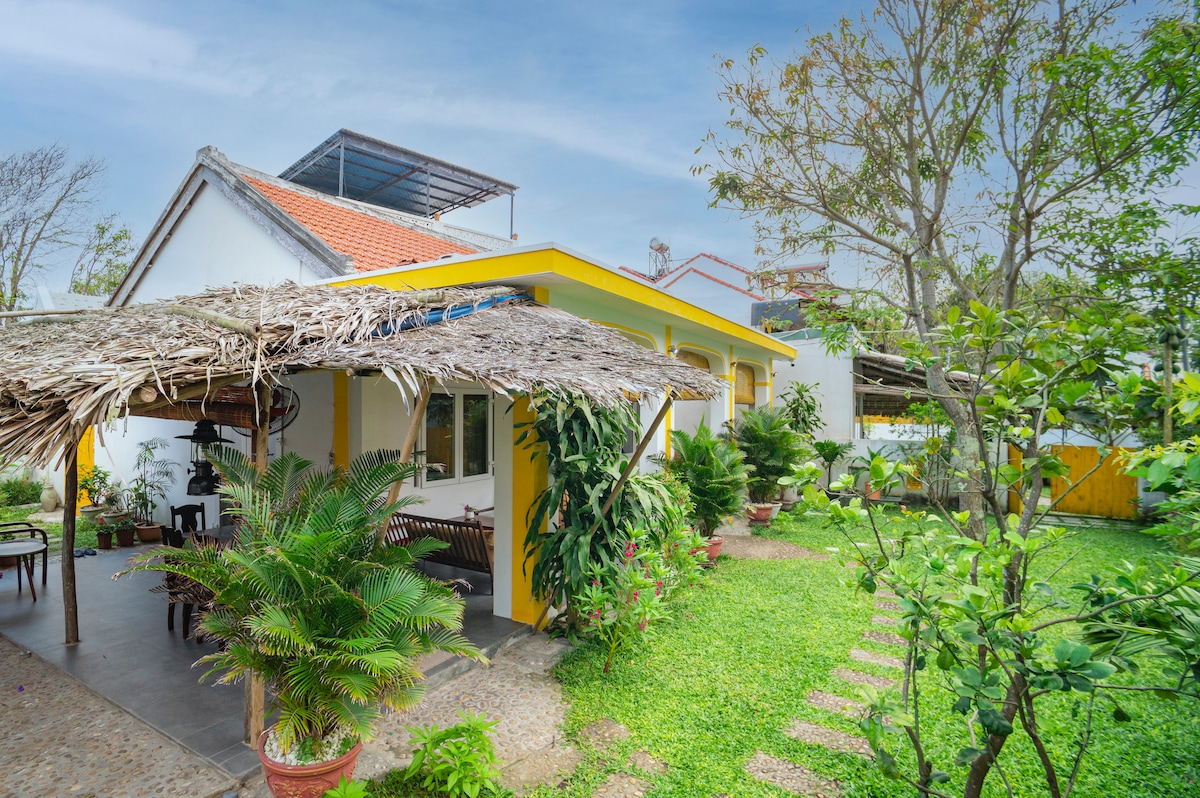 The bungalow features a thatched-roof patio that provides shaded seating, surrounded by greenery and potted plants. A vibrant yellow trim highlights the exterior walls of the home, while a pathway of stepping stones leads through the lush garden.