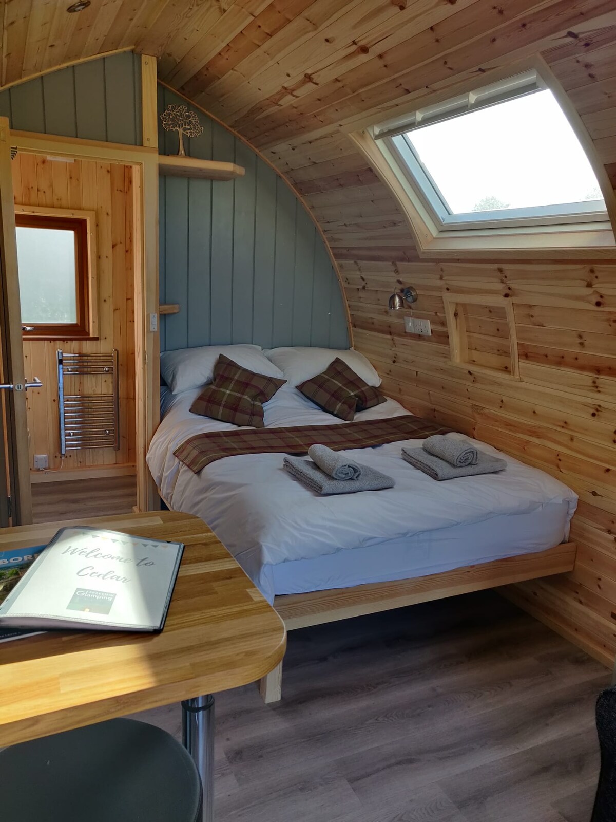 A wooden interior features a double bed with plaid pillows and neatly folded towels on the covers. A window allows natural light to enter, while a small table is visible in the foreground.