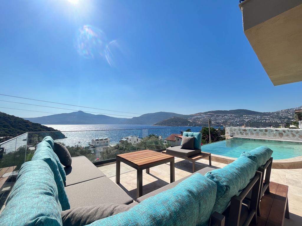 A serene outdoor seating area features turquoise cushions on modern furniture, positioned to offer stunning views of the Kışla bay. The infinity pool creates an illusion of merging with the sea, under a bright blue sky illuminated by sunlight.