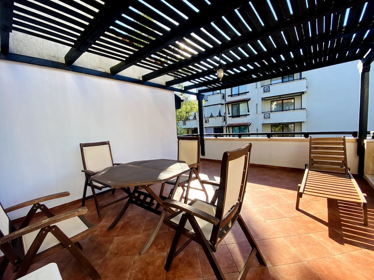 A spacious terrace is featured, furnished with a round wooden table and several wooden chairs. Sun loungers are placed nearby, and the area is shaded by a wooden trellis. The surrounding walls are painted white, providing a light contrast to the terracotta tiled floor.