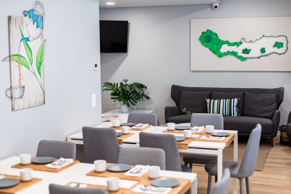 A dining area is presented with several wooden tables set for meals, featuring neutral-toned plates and cups. A cozy seating area with a dark gray sofa is visible, accented by colorful cushions. Modern artwork adorns the walls, enhancing the contemporary decor.