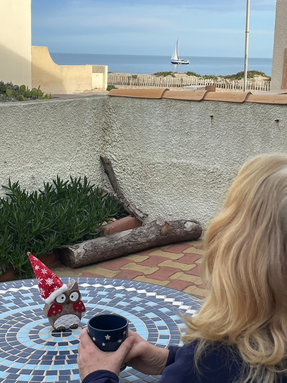 A terrace table decorated with a festive ornament is visible. In the background, the ocean can be seen, with a sailboat sailing in the distance. The setting suggests a peaceful atmosphere, with greenery and stone walls framing the area.