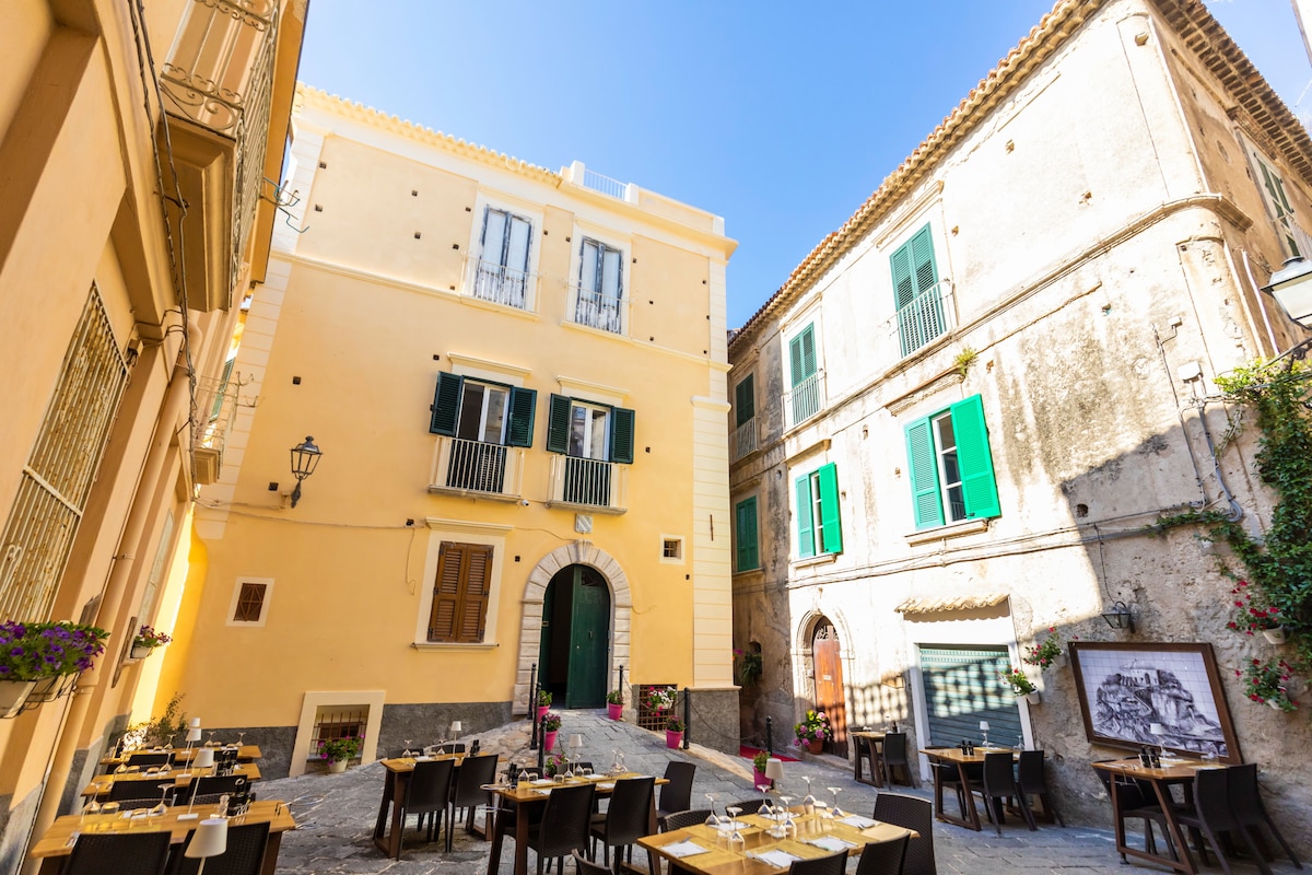 A charming plaza is shown, featuring a historic yellow building with a large arched entrance and multiple windows. Surrounding restaurants have outdoor seating with elegant wooden tables and chairs. Lush flowers accentuate the space, while vibrant green shutters provide contrast against the warm facade.