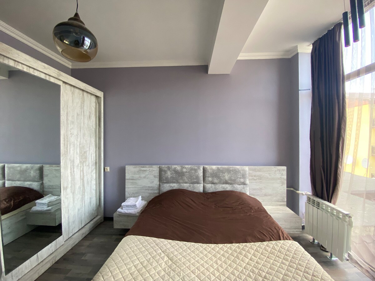 A spacious bedroom features a large bed with a brown coverlet against a muted purple wall. Natural light enters through a window draped with dark curtains. A mirrored wardrobe reflects the room's layout, while neatly arranged towels are displayed on a nearby surface.