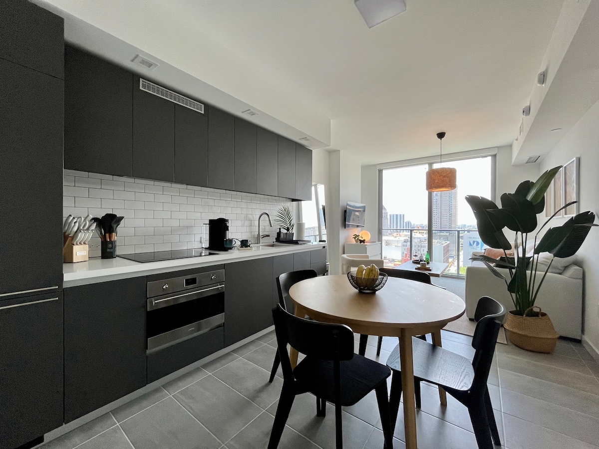 A modern kitchen featuring sleek dark cabinetry and a white countertop. Stainless steel appliances are visible, including an oven and stove. A round dining table with four black chairs is positioned in front of a large window, offering city views. A potted plant adds a touch of greenery.