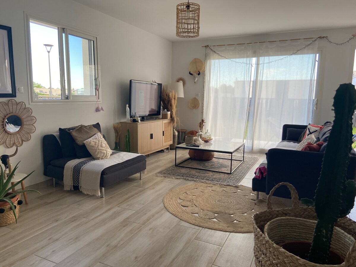 A spacious living area features large windows allowing natural light to fill the room. A black coffee table is centrally located, surrounded by comfortable seating arrangements. Decorative plants and handmade items are visible, enhancing the inviting ambiance of the space.