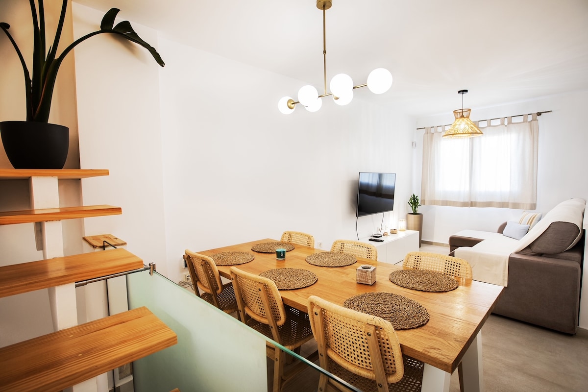 A bright and airy living space features a dining table with woven placemats, surrounded by stylish chairs. A comfortable sofa is positioned nearby, complemented by a soft rug. Natural light flows through a window adorned with light curtains, enhancing the warm ambiance.