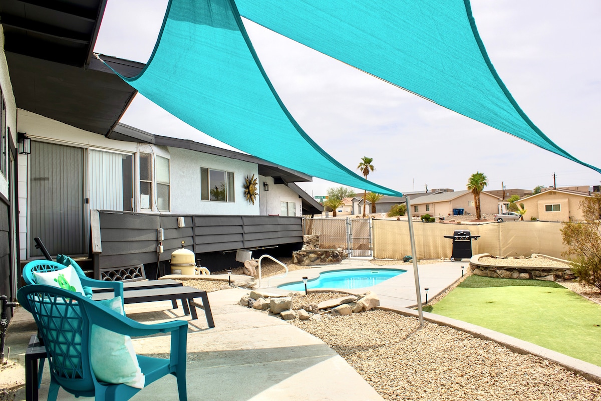 A shaded outdoor area features blue canopies providing comfort beside the sparkling pool. Colorful lounge chairs are positioned to enhance relaxation, while a grassy putting green is visible in the garden. The scene is framed by the clean lines of the renovated property.