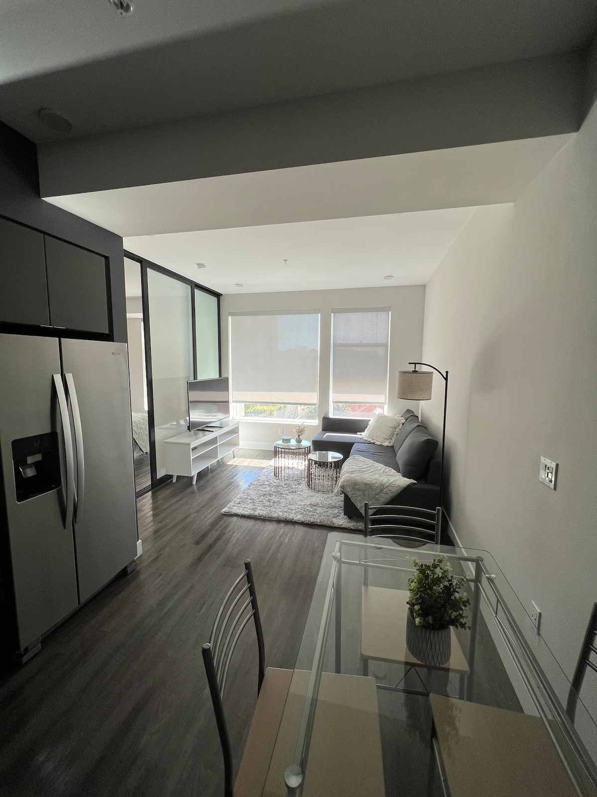 A bright living space showcases a comfortable sofa, a coffee table, and a television stand with a TV. Large windows allow natural light to fill the area, while a dining table with chairs is positioned nearby. The kitchen is visible, featuring modern appliances.