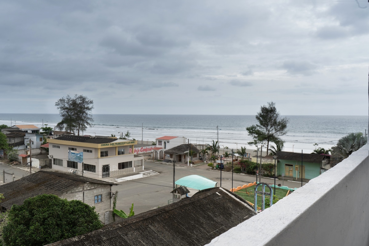 A view of a tranquil beach scene is presented, with a wide shoreline visible beyond a cluster of buildings. The ocean gently meets the sand under a cloudy sky, while a few palm trees and distant structures are lined along the beachfront.