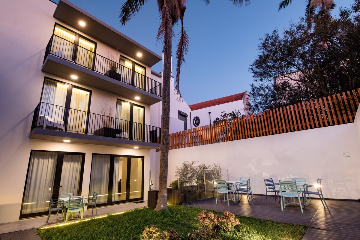 The exterior features a two-story building with balconies overlooking a landscaped area. Small tables and chairs are arranged on the patio, surrounded by greenery. Soft outdoor lighting casts a gentle glow as dusk approaches, creating an inviting setting.