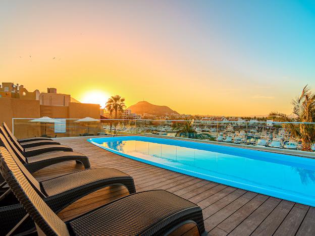 A serene rooftop pool area offers lounge chairs positioned along the edge. The pool reflects the vibrant hues of a sunset, with mountains and the marina visible in the background. Umbrellas are provided for shade, enhancing the inviting outdoor space.