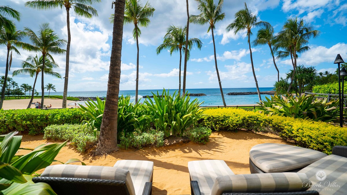 Comfortable lounge chairs are located under swaying palm trees, facing the ocean. Lush green plants and sandy ground surround the seating area, offering a scenic view of the sparkling turquoise water and blue sky dotted with clouds.