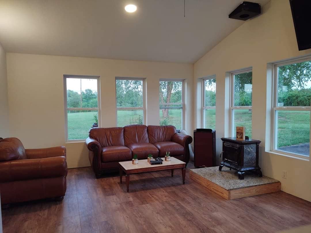 A sunlit sunroom features two leather sofas and a wooden coffee table placed on a laminate floor. Large windows allow natural light to enter, providing views of the surrounding green landscape. A small wood stove is positioned on a stone platform, adding warmth to the space.