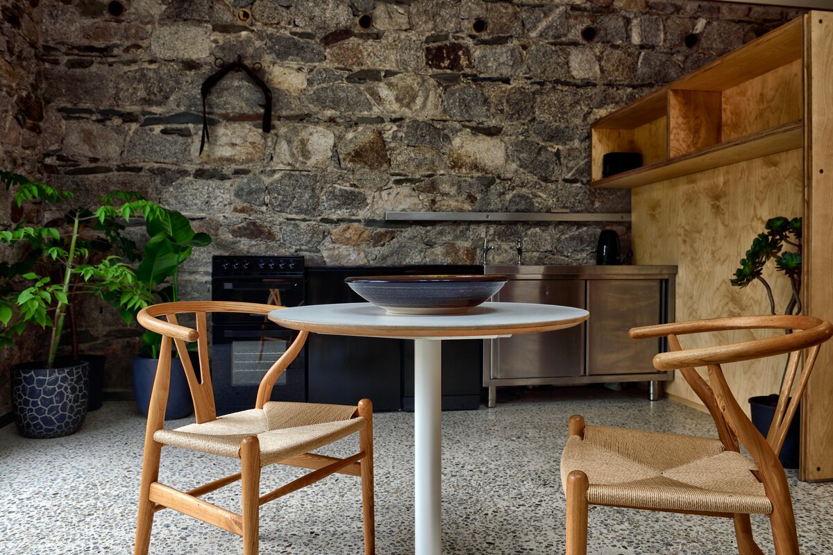 A compact dining area features a round white table surrounded by two wooden chairs with woven seats. Stone walls provide a rustic backdrop, while a kitchenette equipped with stainless steel appliances is visible in the background. Potted plants add a touch of greenery to the space.