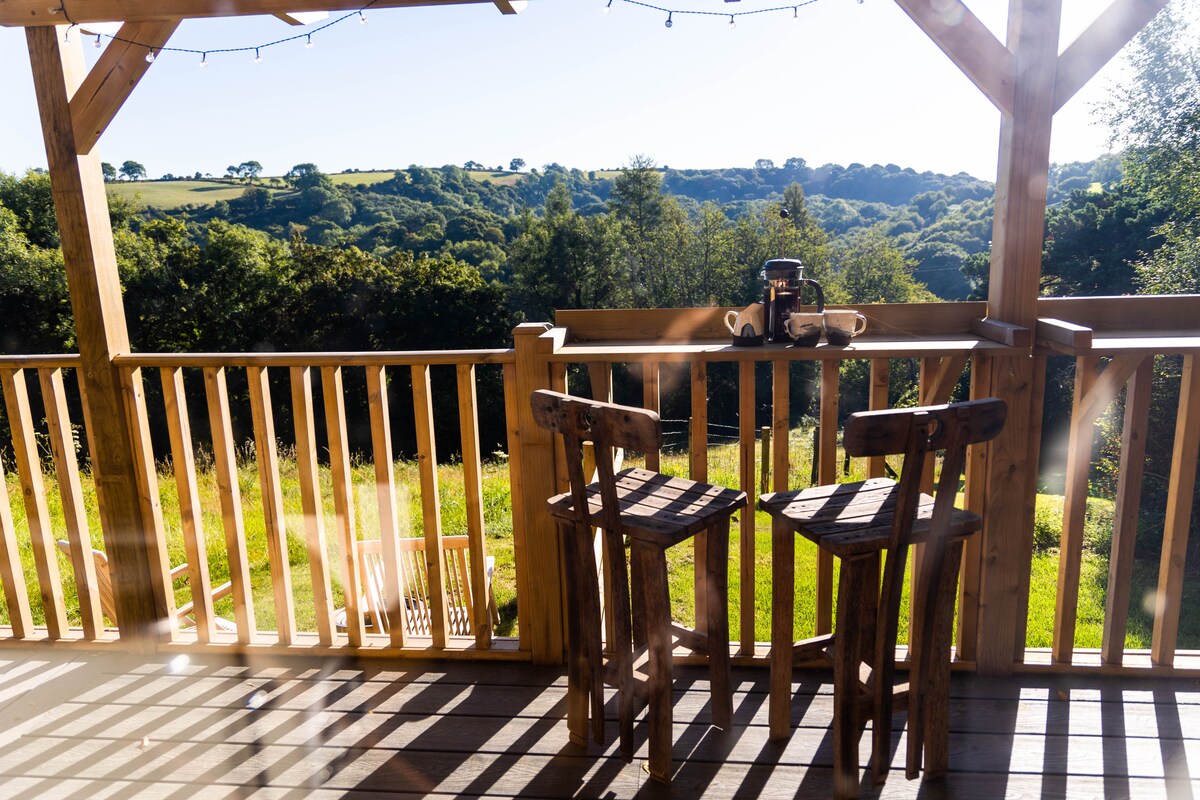 A wooden deck features two rustic chairs positioned next to a small table. The scene is framed by a railing, offering a view of lush green hills and trees in the distance, suggesting a peaceful outdoor setting.