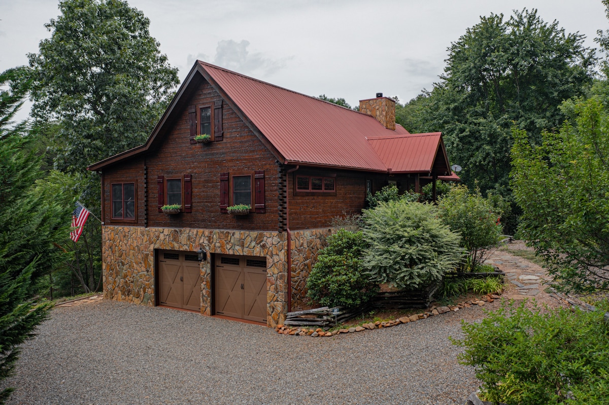 The exterior of a rustic cabin features a combination of wood and stone, with a red metal roof. A gravel driveway leads to the double garage doors, surrounded by well-maintained landscaping and shrubs. American flags adorn the front, enhancing the welcoming appearance.