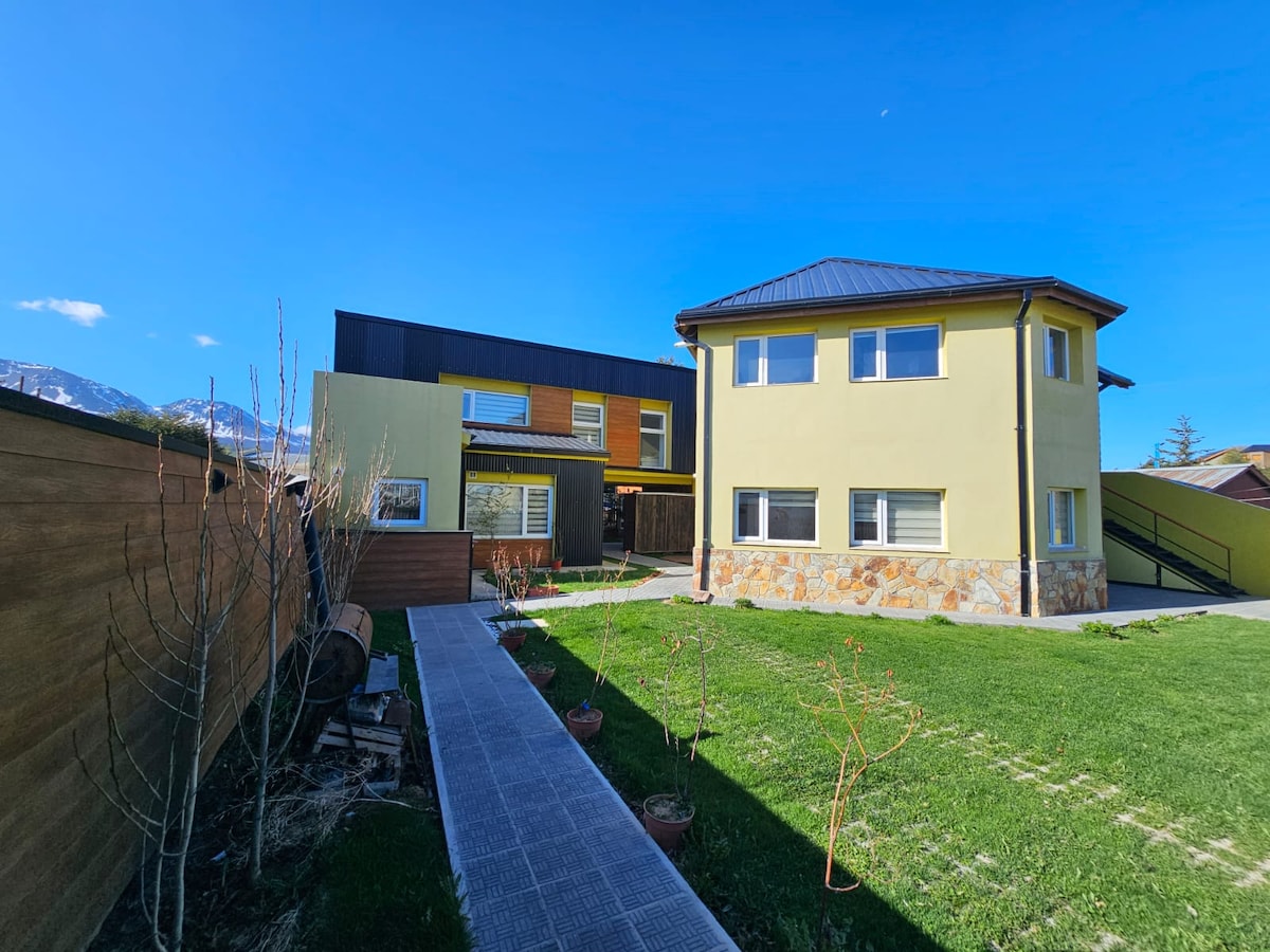 A two-story building featuring a combination of stone and yellow walls, surrounded by a well-kept green lawn. A pathway leads through the garden, while clear skies and distant mountains are visible in the background.