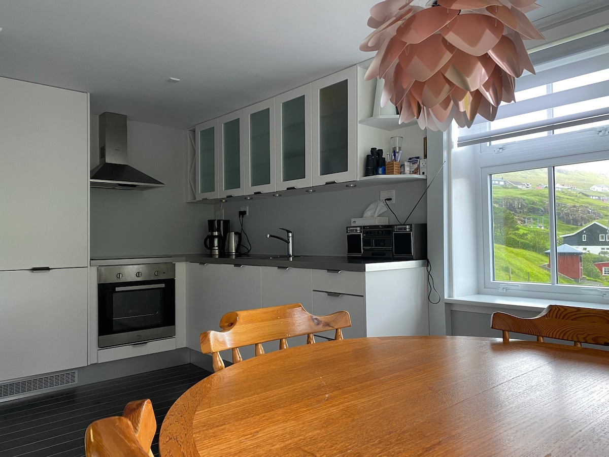 A modern kitchen is seen, featuring sleek white cabinetry and a round wooden table with several chairs surrounding it. A large window allows natural light to illuminate the space, showcasing the view of the surrounding landscape. Appliances include a stovetop, oven, and coffee maker.