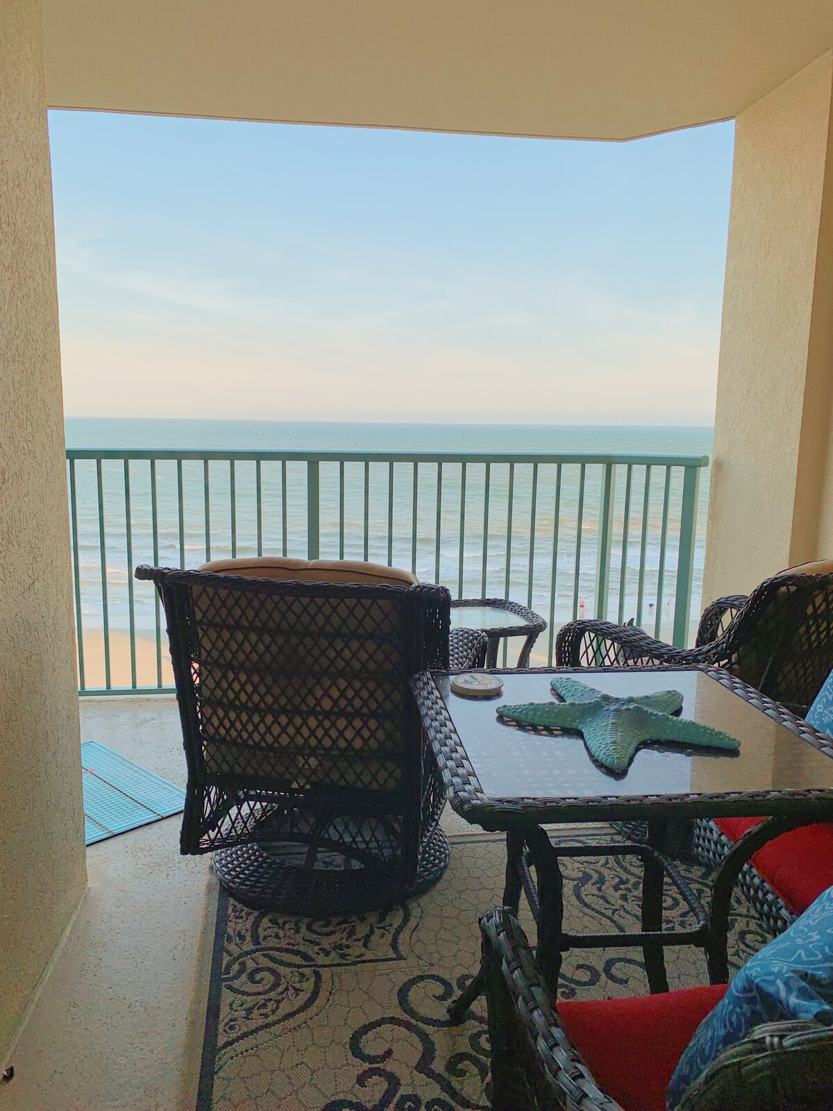 A balcony area features two comfortable wicker chairs and a small table. The space overlooks the ocean, with a railing providing safety and unobstructed views of the beach. The calming sound of waves can be heard from this inviting outdoor retreat.
