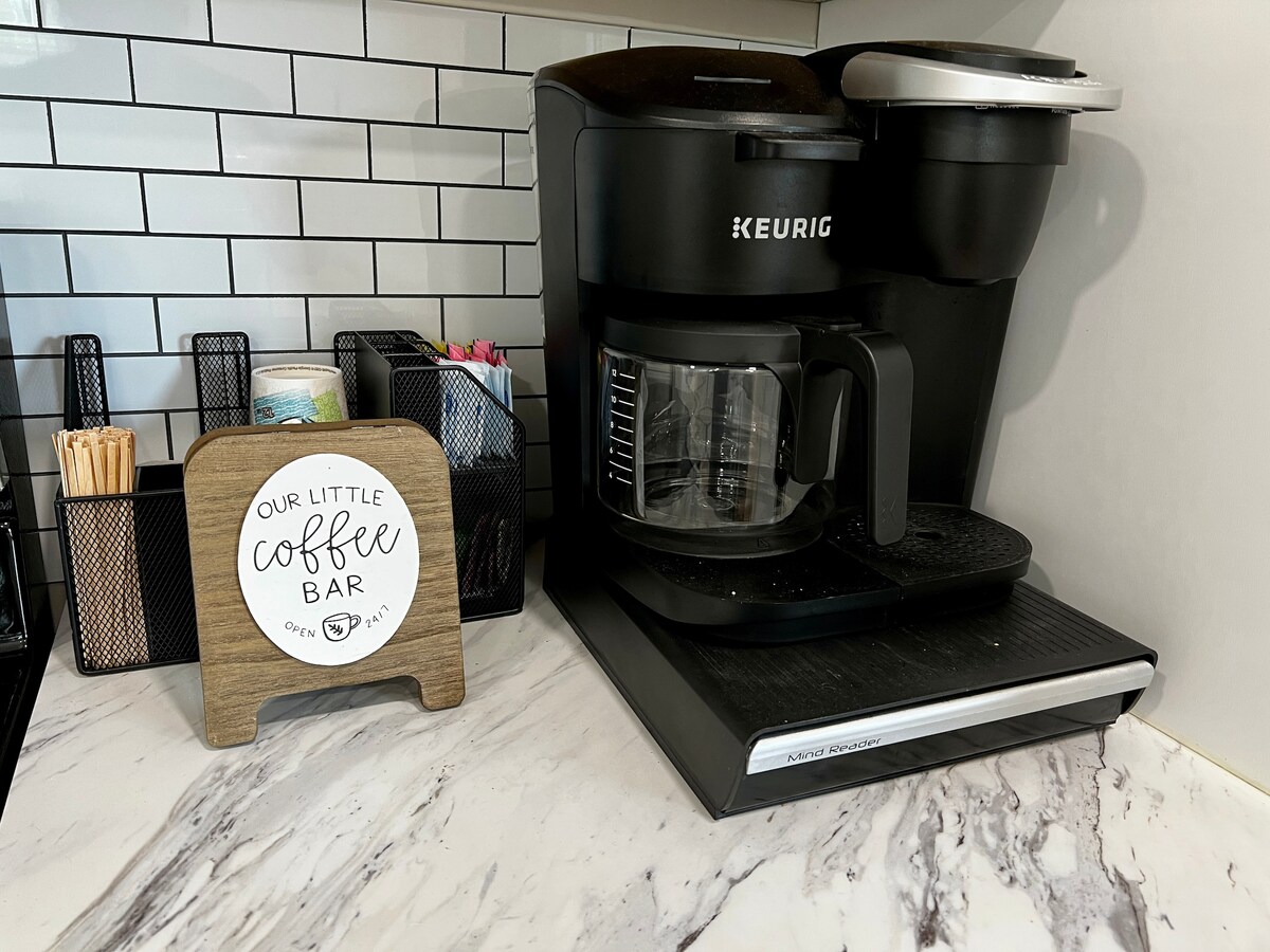 A neatly organized coffee bar features a Keurig coffee maker and a clear glass carafe. A wooden sign stating 'Our Little Coffee Bar' is positioned nearby, along with assorted coffee stirrers stored in a metal holder. The marble countertop is highlighted with light and dark swirling patterns.