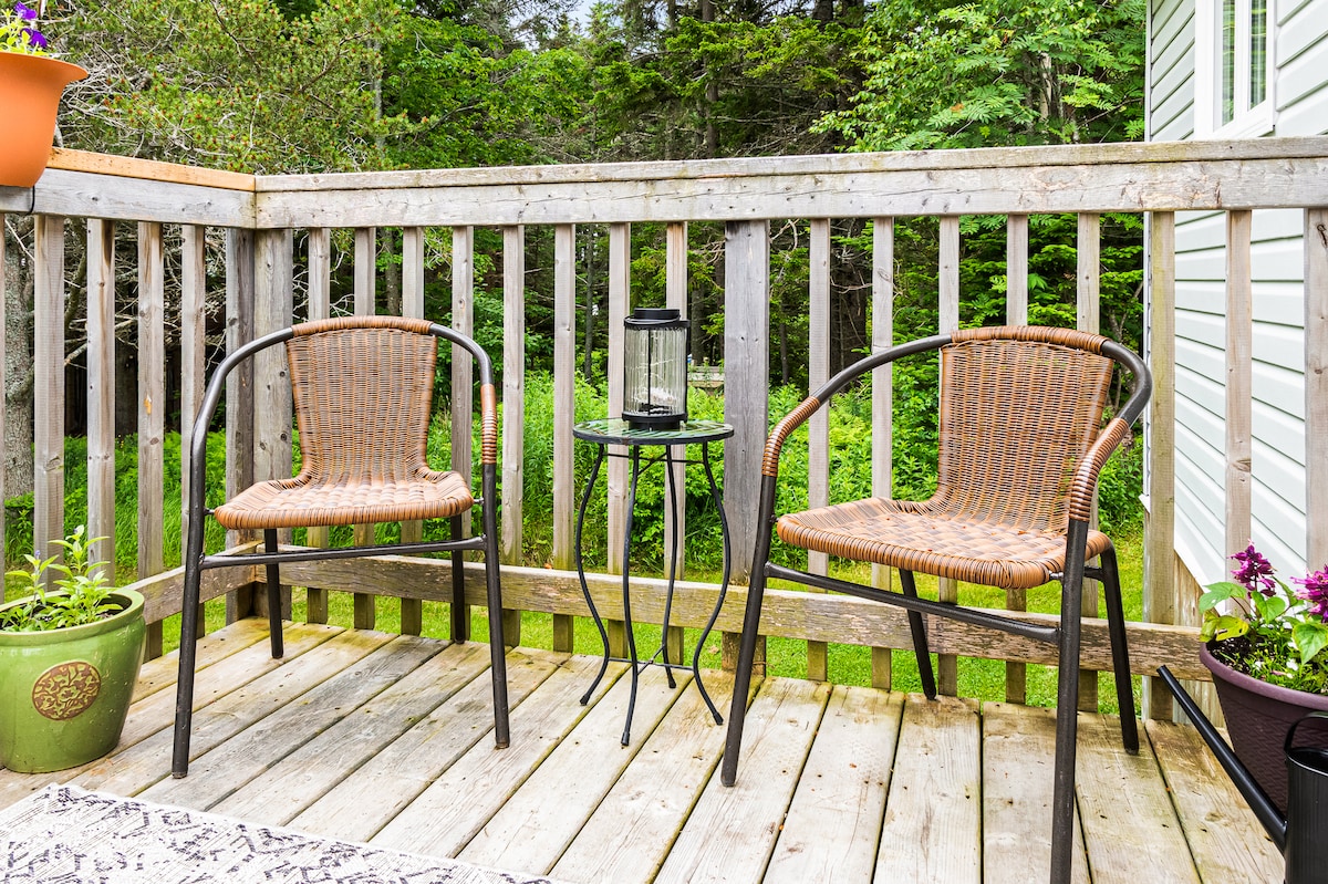 An outdoor space is presented with two woven chairs positioned beside a small black table. A soft lantern sits on the table, complemented by potted plants with green foliage and colorful blooms, surrounded by a natural, wooded backdrop.
