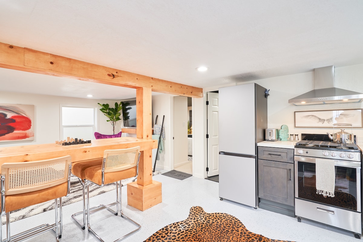The kitchen features modern appliances, including a stainless steel refrigerator and gas stove, complemented by wood beams and bar stools at the counter. A patterned rug adds texture to the space, with a small plant and artwork enhancing the decor.