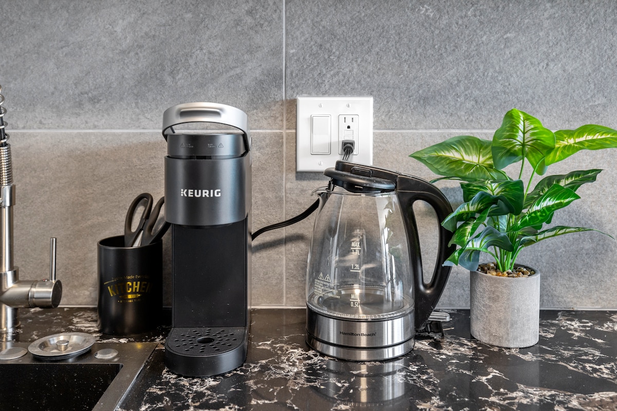 A kitchenette counter showcases a Keurig coffee maker adjacent to an electric kettle. A small potted plant adds a touch of greenery, while the marbled countertop provides a sleek backdrop. Nearby, a mug holder and power outlet are visible.