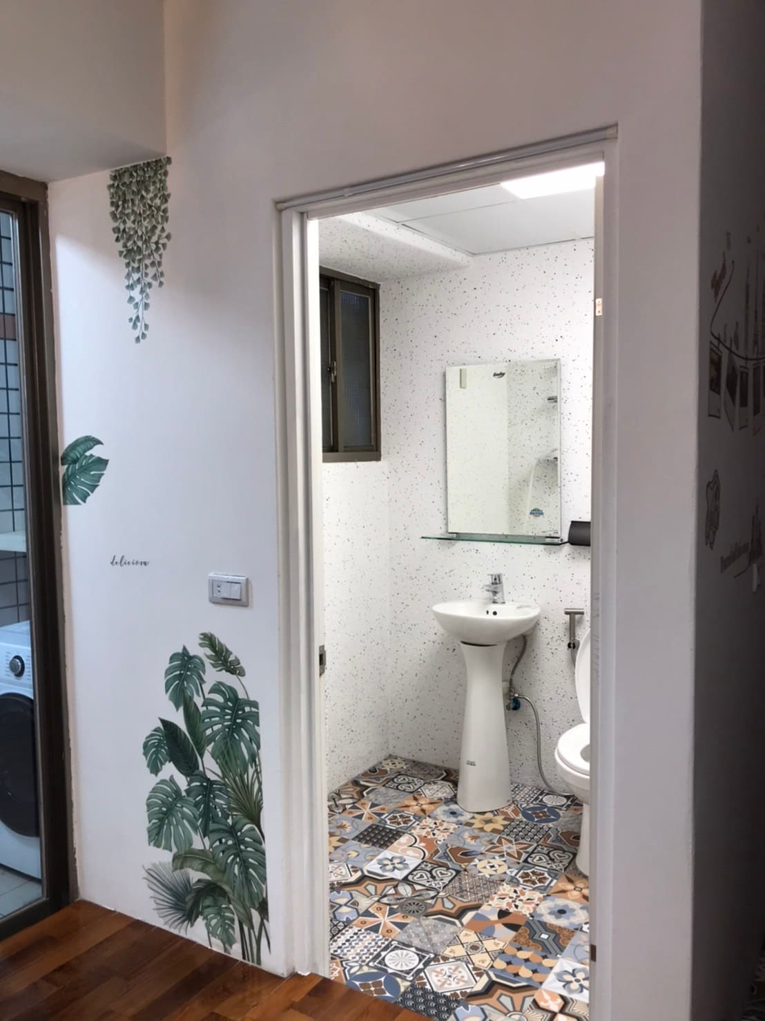 A clean bathroom entrance is framed by patterned tiles and decorative wall plants. Inside, a modern sink and mirror are visible alongside a toilet. The space features bright lighting, creating a fresh and tidy atmosphere.