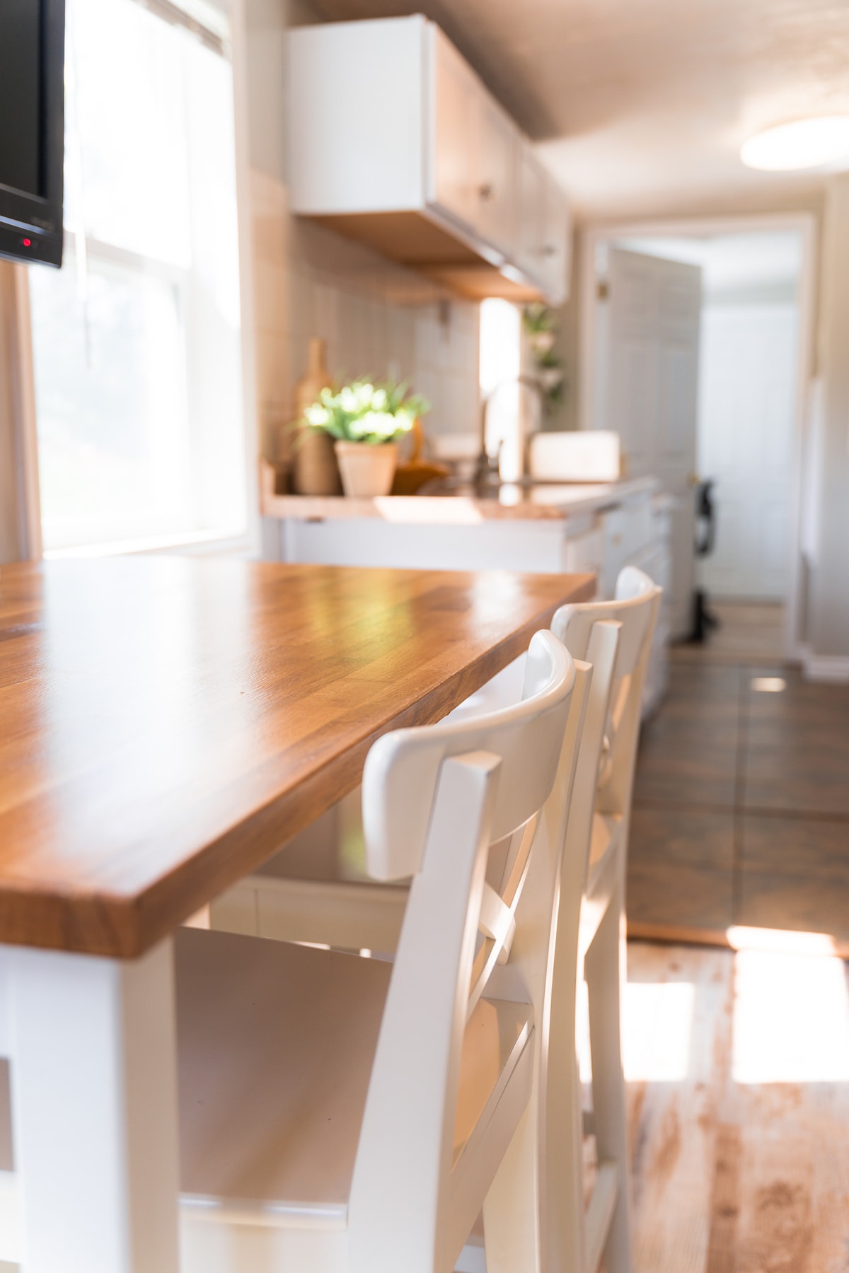 A well-lit kitchen area is showcased, featuring a wooden bar counter with two white chairs. Above the counter, cabinets provide storage, and potted plants bring a touch of greenery to the space. Natural light floods in through the windows, enhancing the clean and functional design.