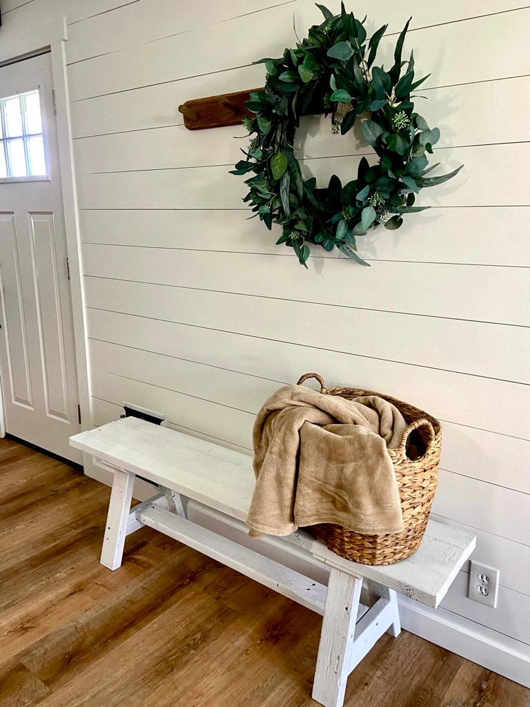 A welcoming entryway is presented, featuring a rustic white bench beside a door. A circular greenery wreath is hung on the wall above, complemented by a natural woven basket containing a folded blanket, enhancing the cozy atmosphere.