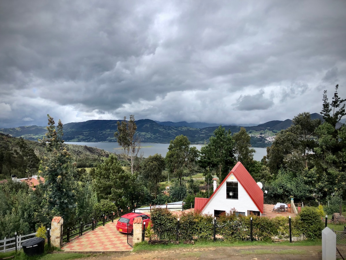 A scenic view captures a chalet with a red roof surrounded by greenery. A serene lake reflects the overcast sky, with mountains in the distance. The patterned driveway leads to the entrance, providing a welcoming atmosphere against a backdrop of nature.
