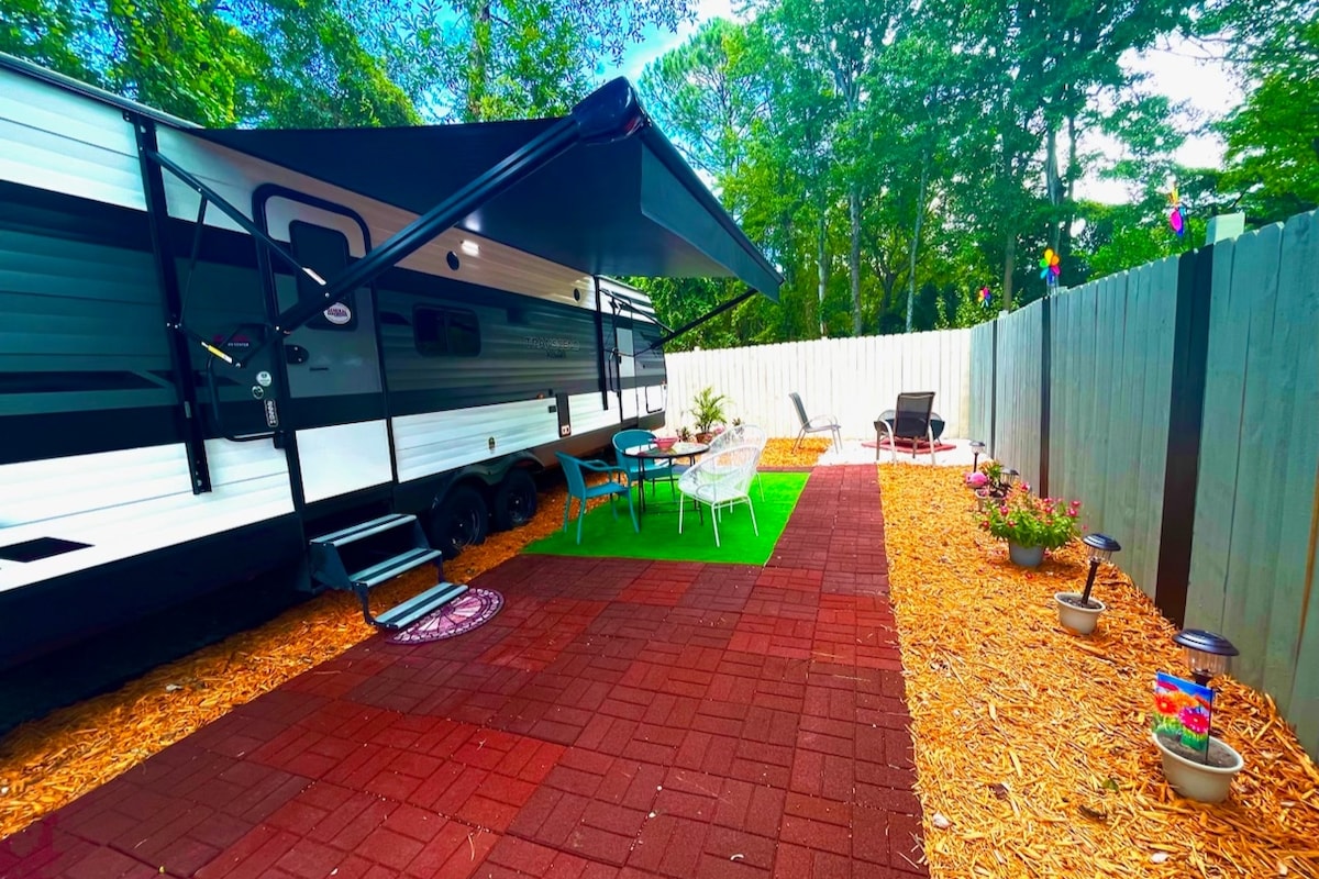 An outdoor seating area is presented next to a parked RV, featuring a green patch of grass. A cozy table is surrounded by four chairs, while two additional lounge chairs are positioned nearby. Colorful flower pots add vibrancy to the space, bordered by a privacy fence.