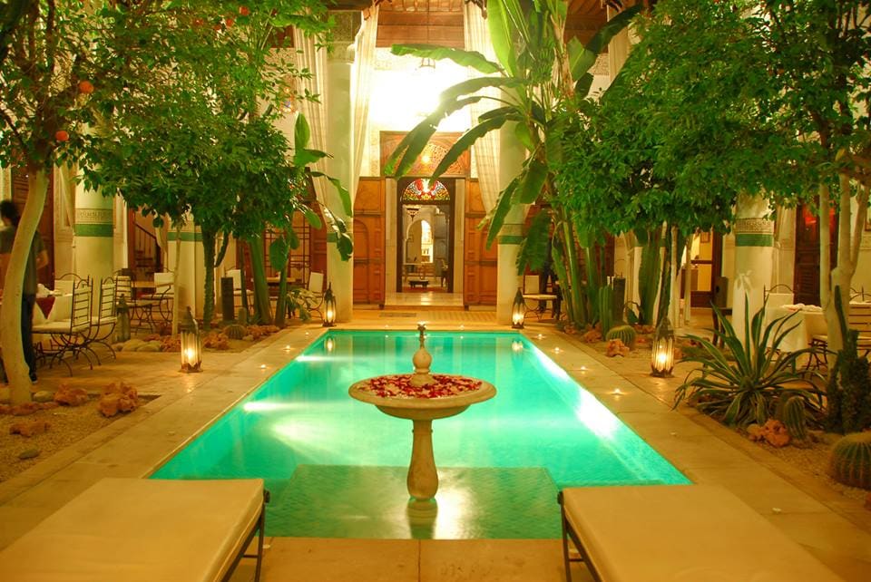 A serene courtyard is highlighted by a heated swimming pool surrounded by lush greenery and decorative plants. Soft lighting enhances the tranquil atmosphere, while comfortable loungers are positioned along the water's edge. The space is designed for relaxation and reflection in a traditional Moroccan setting.