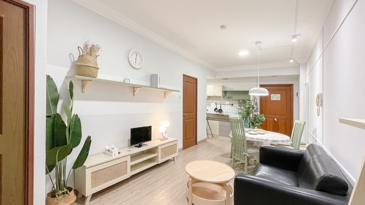 The living area features a comfortable black sofa paired with a light wooden coffee table. A small dining space is visible, with green chairs surrounding a round table. Decorative plants are placed near the wall, and a TV is positioned on a shelf beneath a clock.