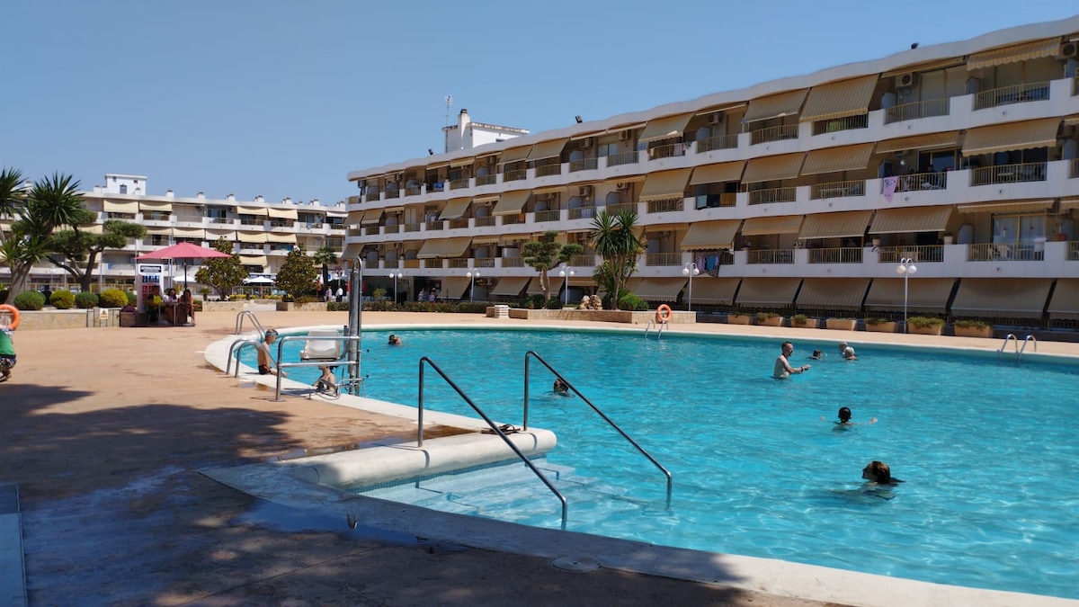 A large swimming pool is surrounded by sun loungers and palm trees, offering a recreational area for all ages. The pool is framed by a multi-story building with numerous balconies, enhancing the vibrant atmosphere. Sunbathers and swimmers can be seen enjoying the water.
