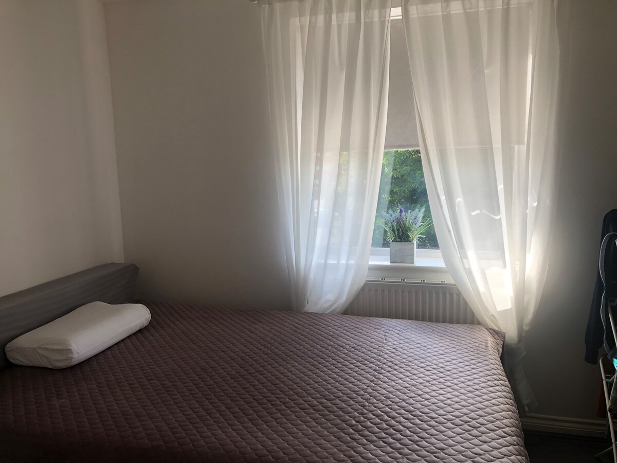A clean and simple sleeping area features a spacious bed with a textured quilt in muted tones. Natural light filters through sheer white curtains, illuminating the room. A small potted plant is visible on the windowsill, adding a touch of greenery.