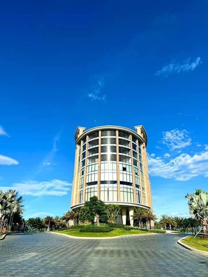 A modern building is set against a clear blue sky, featuring large glass panels that reflect the surroundings. Lush green landscaping and palm trees are visible at the base, leading to a circular driveway that surrounds the structure.