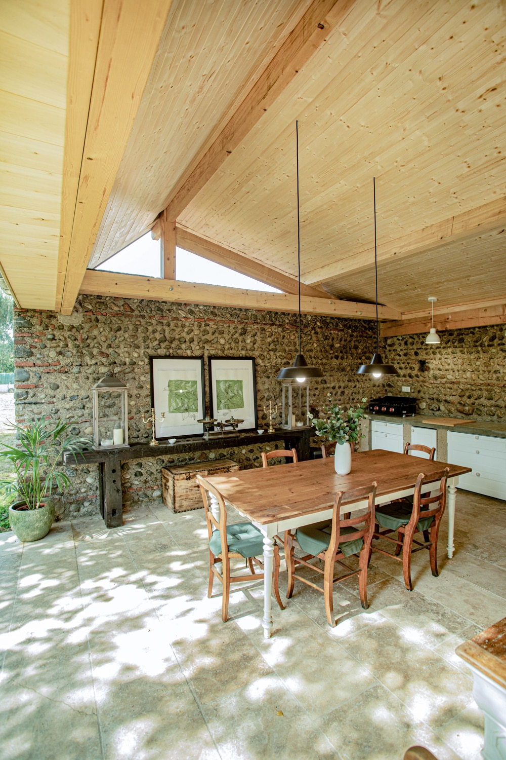 A spacious dining area features a wooden table surrounded by six chairs, complemented by a rustic sideboard. Exposed wooden beams enhance the ceiling, while large windows allow natural light to fill the space. Lush greenery is positioned near the window, bringing a sense of calm.