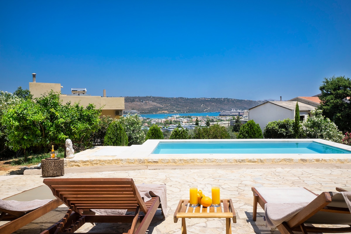 A serene outdoor area is highlighted by a private pool and views of the sea in the background. Sun loungers are positioned nearby on a stone patio, while lush greenery frames the space, adding to the tranquil ambiance.