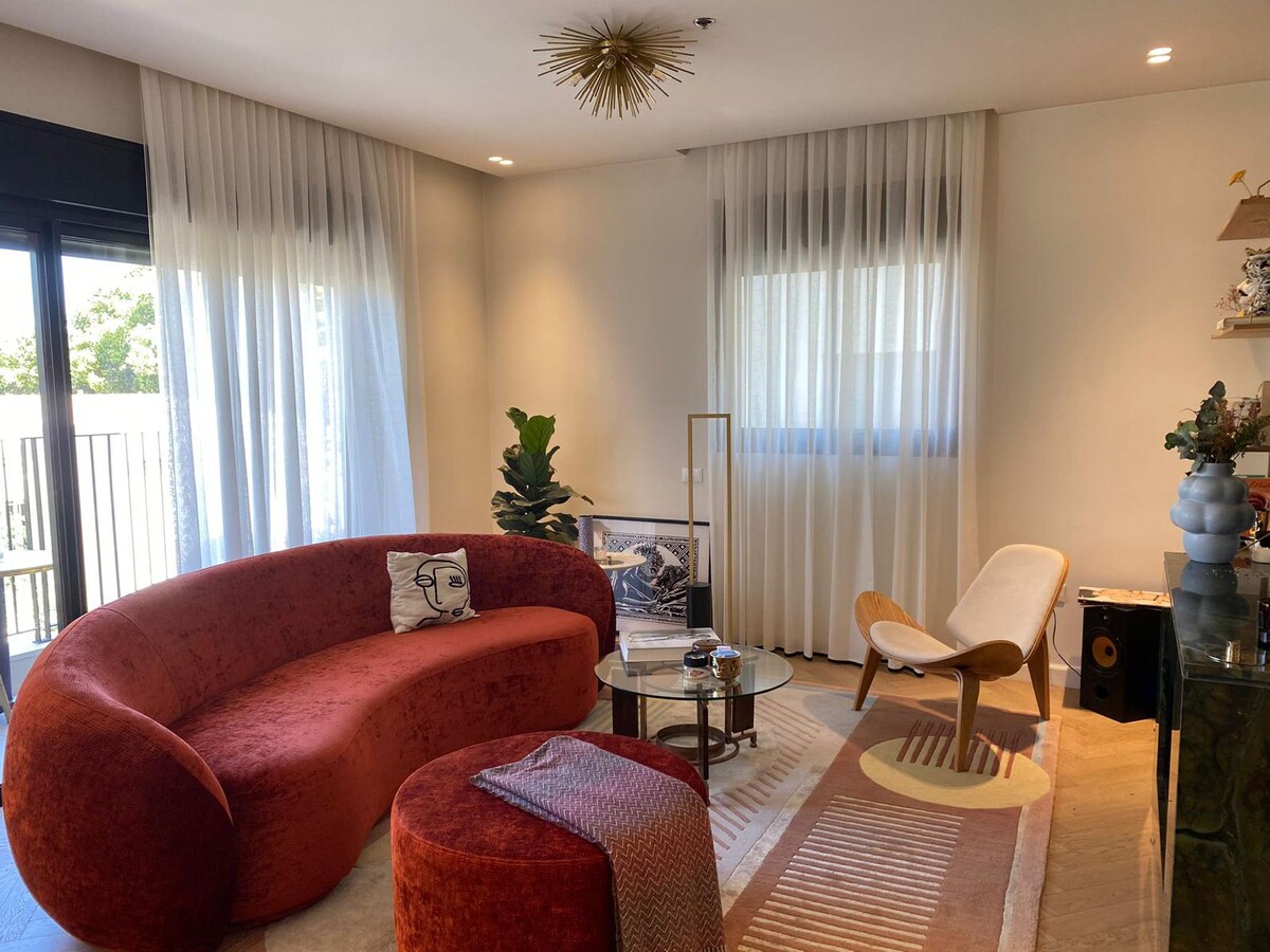 A spacious living room features a curved red sofa and a light-colored accent chair. Natural light fills the area through large windows with sheer curtains. A round glass coffee table is positioned on a textured area rug, complemented by decorative plants and a modern light fixture overhead.